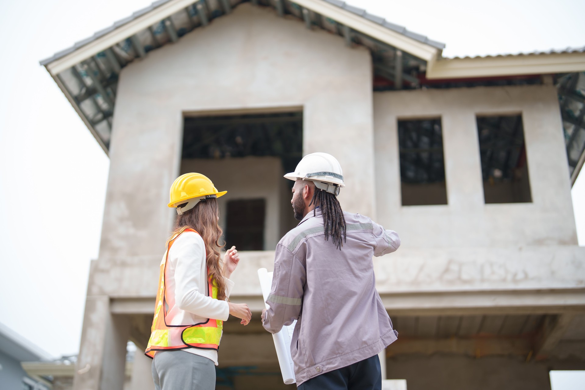 Back view of construction workers standing in front of unfinished house, male and female professionals in safety gear discussing progress and pointing towards building structure, showing teamwork and supervision on site